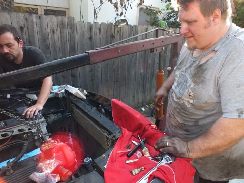 2003 Ford Crown Victoria - grease-streaked man sorts hand tools laid on a red cloth at the front of a vehicle as another works at the radiator.