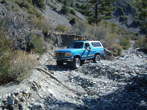 1988 Ford Bronco - A blue Ford Bronco navigates a rough, rocky terrain in a mountainous landscape.