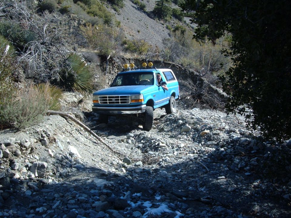 Blue Ford Bronco with roof-mounted lights driving on a rocky, dry creek bed
