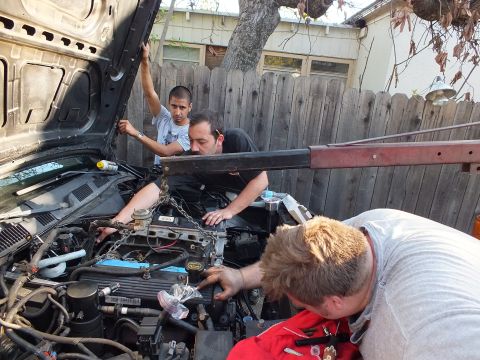 2003 Ford Crown Victoria - Three men reach into the engine bay of a hood-up vehicle in a fenced backyard driveway.