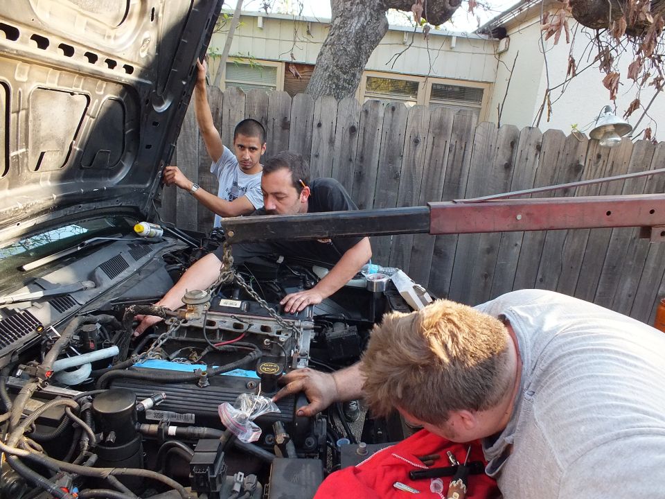 Three men work over open engine bay of SUV in fenced driveway