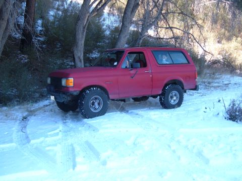 1988 Ford Bronco - A red SUV, likely a 1987 model, navigates a snowy terrain with trees in the background.