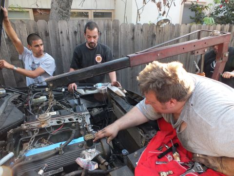 2003 Ford Crown Victoria - group lowers an engine into a vehicle bay, one man reaching to a valve-cover cap under the hoist beam.