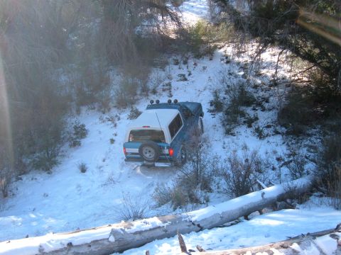 1988 Ford Bronco - A blue SUV with a white camper shell navigates a snow-covered trail, with a fallen log in the foreground.