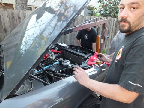 2003 Ford Crown Victoria - bearded man in a shop T-shirt stands at the open hood of a gray vehicle while another works in the bay behind.