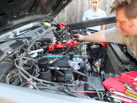 2003 Ford Crown Victoria - worker reaches deep into a wiring-filled engine bay showing a Ford valve cover, with red cloths and wrenches nearby.