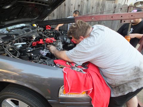 2003 Ford Crown Victoria - man leans across the nose of a gray sedan, working in the engine bay with a red towel over the fender.