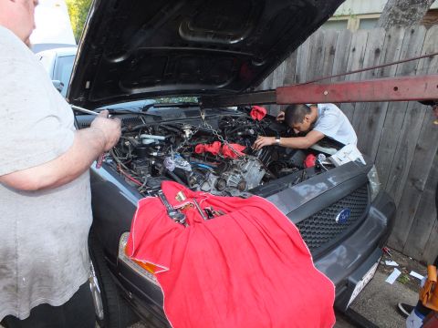 2003 Ford Crown Victoria - Workers reach into the bay of a dark Ford SUV, a red cloth draped over the grille bearing the Ford oval.