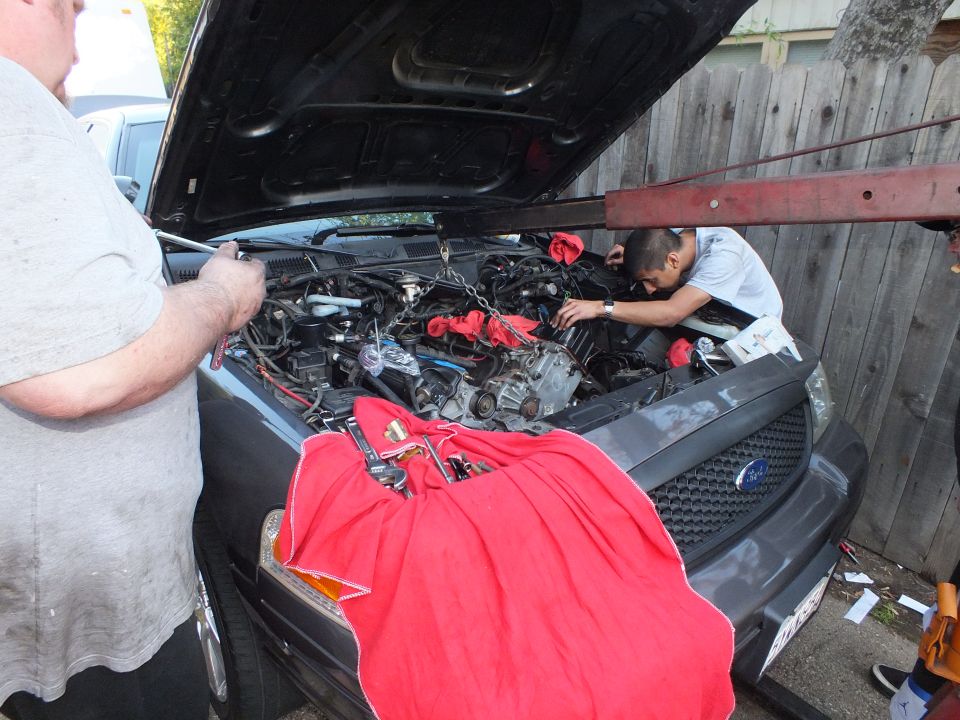 Men work in engine bay of dark Ford SUV with red cloth draped over grille