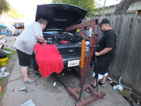 2003 Ford Crown Victoria - Engine crane positioned at the front of a Ford with raised hood, plate 5YWN854, as workers operate the lift outdoors.