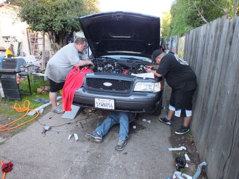 2003 Ford Crown Victoria - Front view of a Ford with hood up, person lying beneath the bumper, two men reaching into the engine bay.