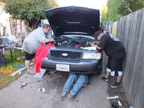 2003 Ford Crown Victoria - Driveway repair on a Ford SUV, hood open, legs visible beneath the front end and tools scattered on the ground.