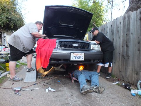 2003 Ford Crown Victoria - Front-end work on a grey Ford against a wood fence, droplight glowing under the bumper near a person's legs.