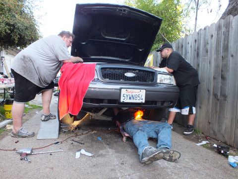 2003 Ford Crown Victoria - Two men lean over the open hood of a grey Ford while another lies beneath the front end with a work light.