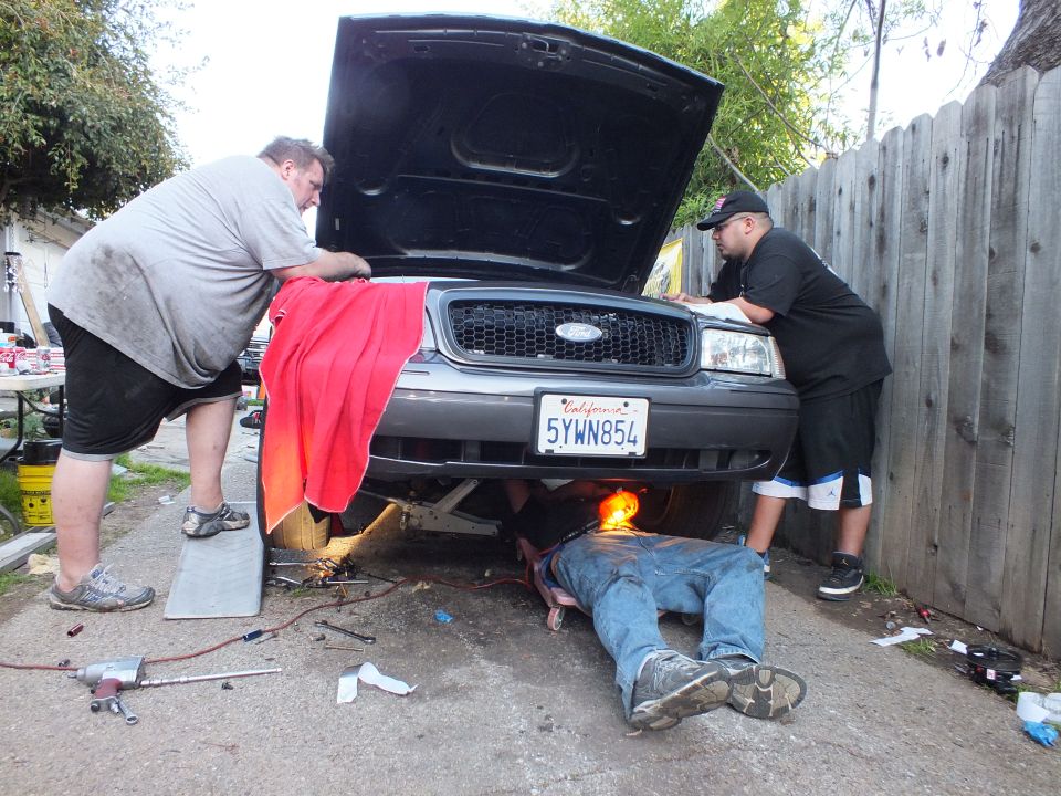Workers service the engine bay of a Ford with California plate 5YWN854 in a fenced driveway