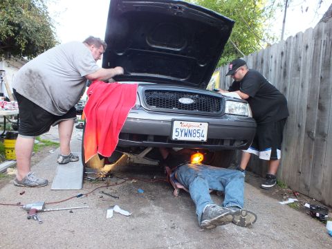 2003 Ford Crown Victoria - Front of a grey Ford with hood open, person under the bumper, two men reaching into the engine compartment.