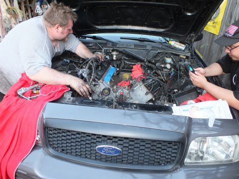 2003 Ford Crown Victoria - Open engine compartment of a grey Ford with blue oval grille badge; one worker leans in, another checks a phone.