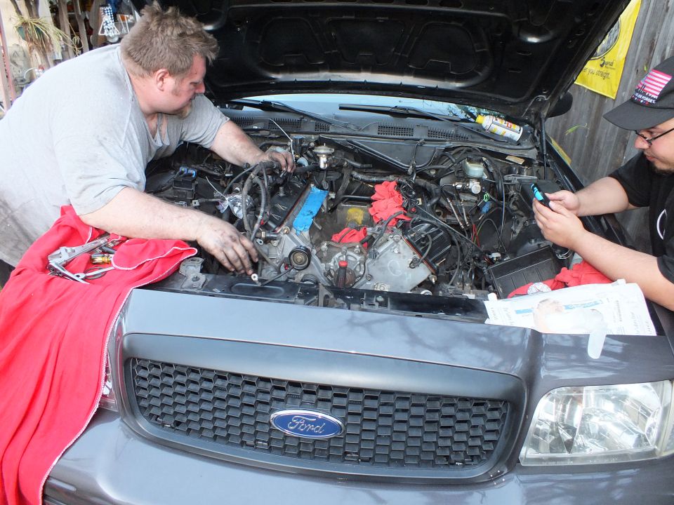 Man reaches into the engine bay of a grey Ford SUV while another holds a phone and paperwork