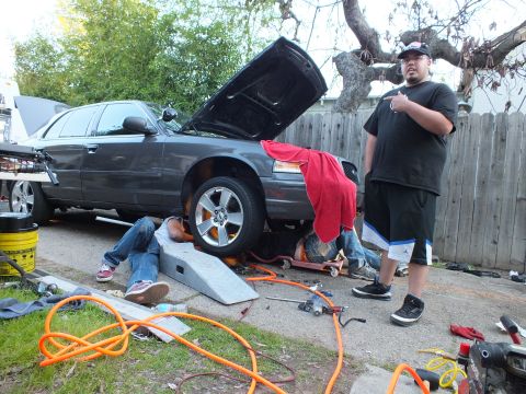 2003 Ford Crown Victoria - Rear-three-quarter view of a dark Ford sedan on a ramp, hood up, two people working underneath outdoors.