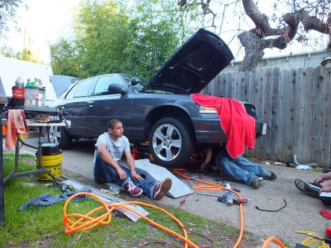 2003 Ford Crown Victoria - Dark sedan raised on a ramp with hood up, orange air hoses on the ground and a seated man beside the front wheel.
