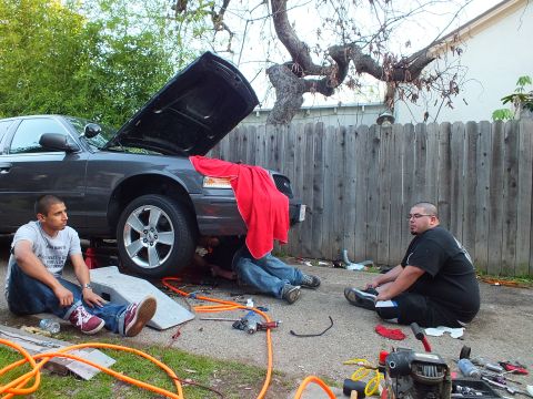 2003 Ford Crown Victoria - Dark sedan on a ramp against a wood fence, two seated workers and scattered tools and hoses in the foreground.
