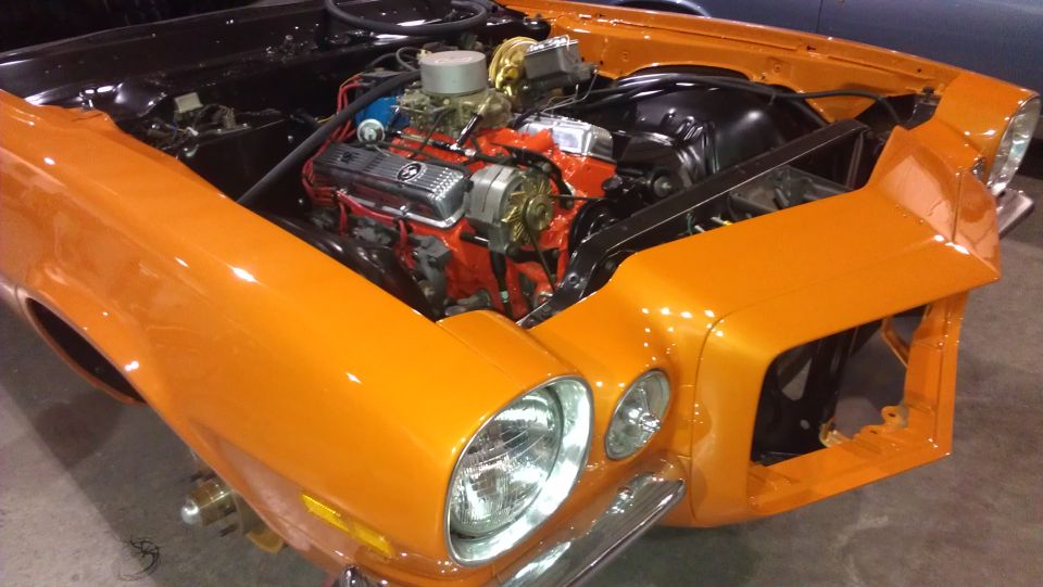 Orange 1970 Chevrolet Camaro front end with hood open, showing engine. Chrome bumper and headlights visible