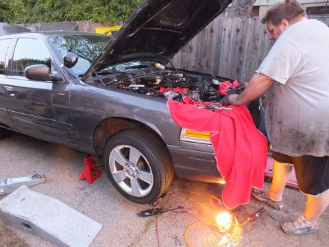 2003 Ford Crown Victoria - Grey sedan raised on a jack stand, hood up, droplight glowing as a man reaches into the engine bay over a red rag.
