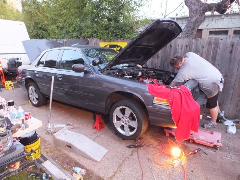 2003 Ford Crown Victoria - Side profile of a grey four-door sedan lifted on stands, hood raised, work light beneath and a creeper on the ground.