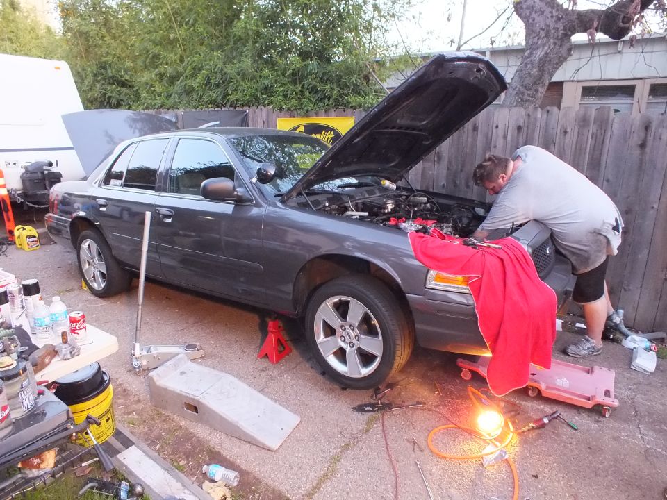 Grey Ford sedan on jack stands with hood open and a man servicing the engine bay