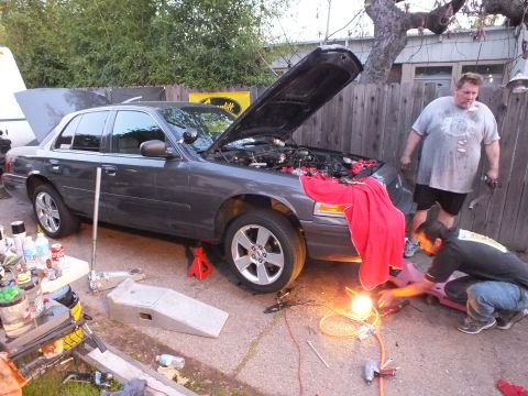2003 Ford Crown Victoria - Grey sedan lifted on stands, one man kneeling with a work light at the front wheel, another standing nearby.