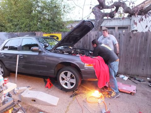 2003 Ford Crown Victoria - Grey four-door sedan on stands, two workers bent over the front fender, droplight illuminating the lower bumper.