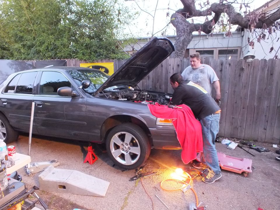 Men work at the front wheel of a grey Ford sedan raised on jack stands with hood open