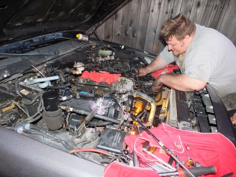 2003 Ford Crown Victoria - Close view of a grey vehicle's engine compartment as a man reaches in, yellow droplight glowing at the fender.
