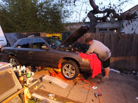 2003 Ford Crown Victoria - Grey four-door sedan lifted on stands against a fence, hood up, a man bent over the front fender at twilight.