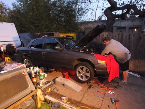 2003 Ford Crown Victoria - Side view of a grey sedan on stands at dusk, red rag over the fender and a man reaching into the engine bay.