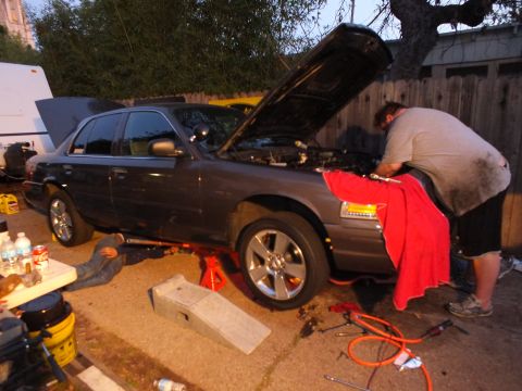 2003 Ford Crown Victoria - Grey four-door sedan lifted on stands at dusk, legs visible beneath and a worker leaning over the front fender.