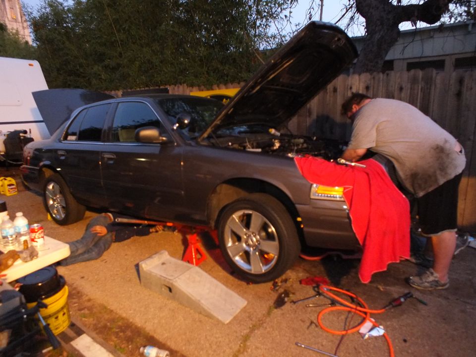 Grey Ford sedan with amber markers on jack stands, hood open, man working at the front end