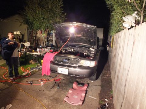 2003 Ford Crown Victoria - Front of a grey Ford at night, hood raised with a hanging work light, man standing at left amid tools and hoses.