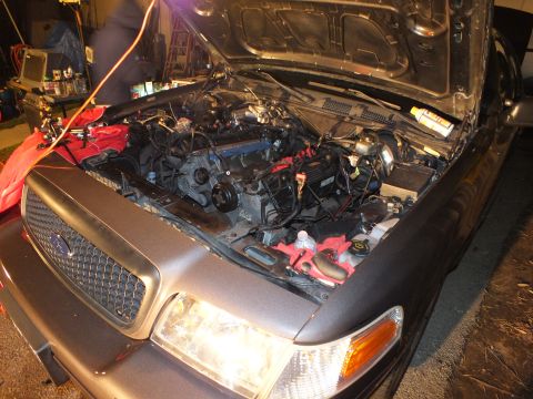 2003 Ford Crown Victoria - Front three-quarter view into a grey Ford's engine bay at night, amber turn lamp lit, blue oval grille badge.