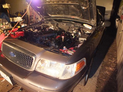 2003 Ford Crown Victoria - Grey Panther-style Ford sedan at night, hood up, honeycomb grille and amber turn lamp lit, engine partly stripped.
