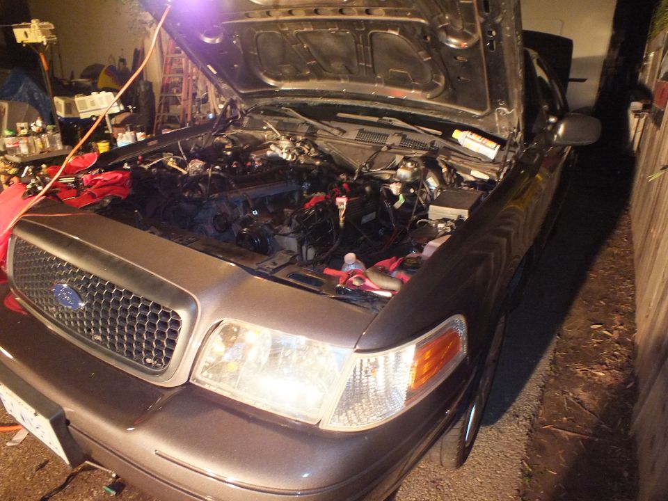 Grey Ford Crown Victoria style sedan with honeycomb grille and open hood under repair at night