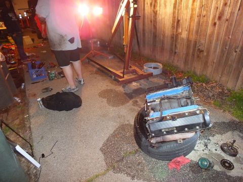 2003 Ford Crown Victoria - Blue-taped V6 cylinder head rests on a tire near an engine stand during a nighttime garage session
