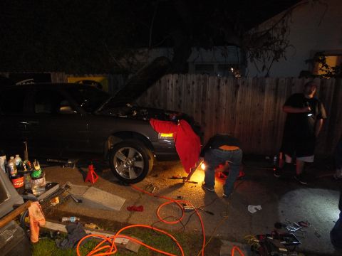 2003 Ford Crown Victoria - Hood-up SUV under a tree at night, a worker crouches over parts amid coiled orange air lines