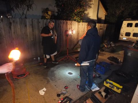 2003 Ford Crown Victoria - Two men talk over a tool spread in a dim backyard, lit by a portable stand light and lantern
