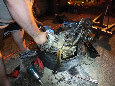 2003 Ford Crown Victoria - Worker pulls a part from the V6 on a tire, a blue cordless impact wrench lying on the pavement