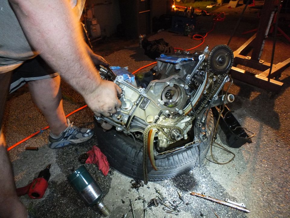 Man lifts a component off a disassembled V6 engine on a tire at night, impact tool on the ground