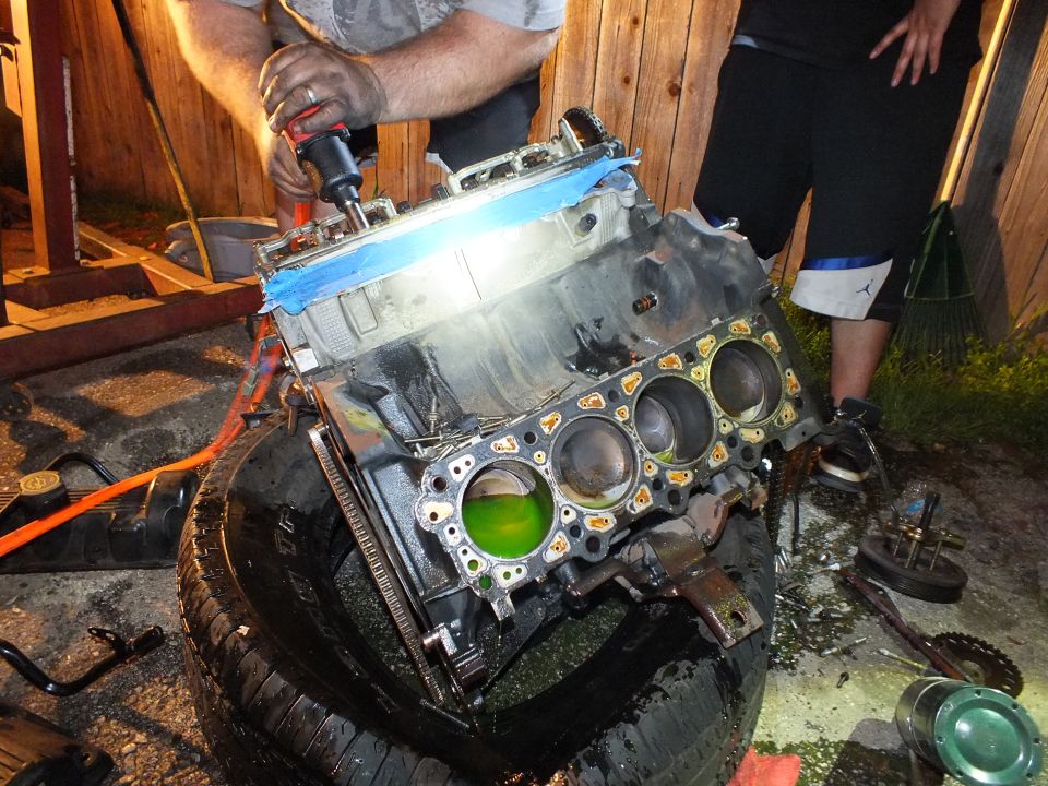 Man uses a cordless drill on an engine block with exposed bores resting on a tire at night