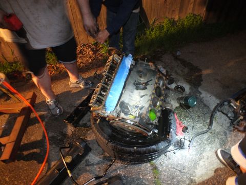 2003 Ford Crown Victoria - Blue-taped V6 rests on a tire while workers handle parts behind it in a dim fenced backyard