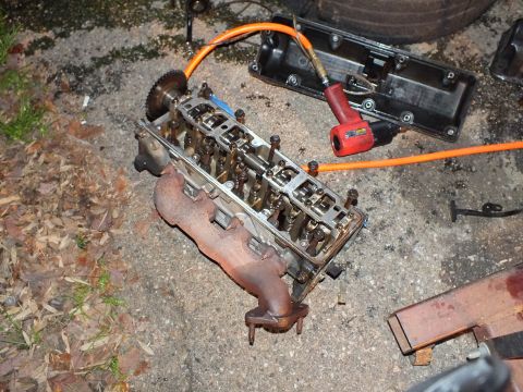 2003 Ford Crown Victoria - Removed cylinder head with valvetrain and a rusty exhaust manifold lie on the ground near a drill
