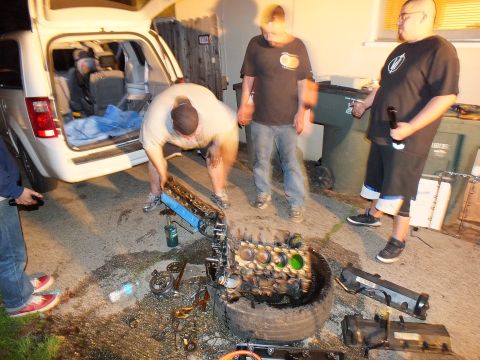2003 Ford Crown Victoria - worker bends over an engine block on a tire while others watch in a nighttime driveway